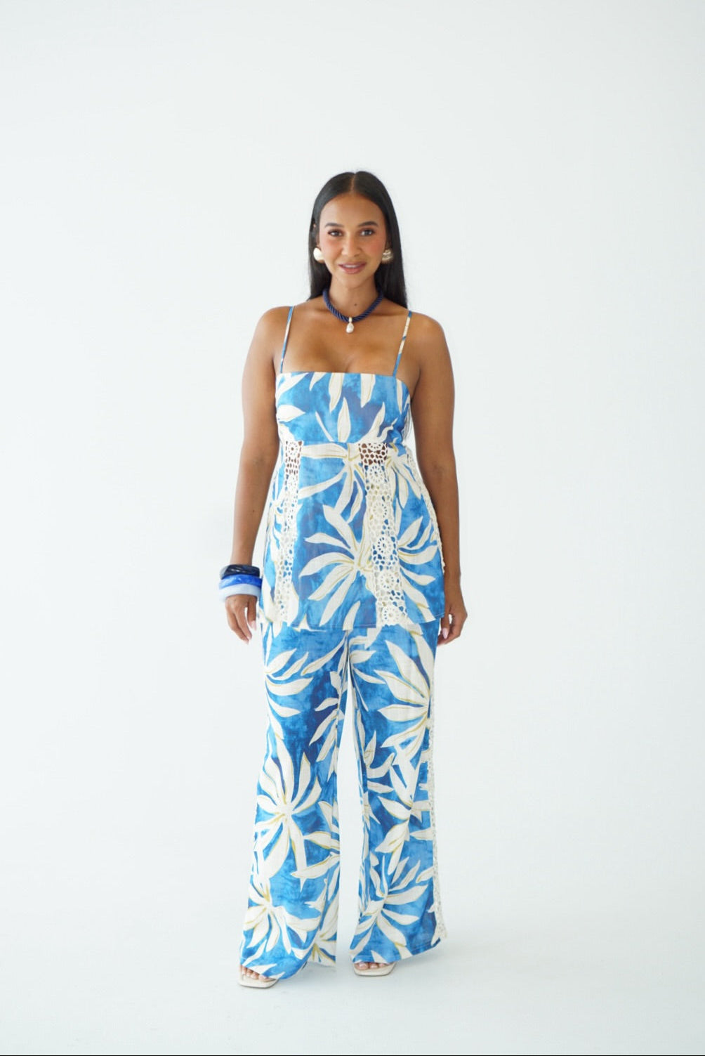 A woman modeling a blue and white tropical floral print sleeveless jumpsuit with a matching belt, standing against a plain white studio background.