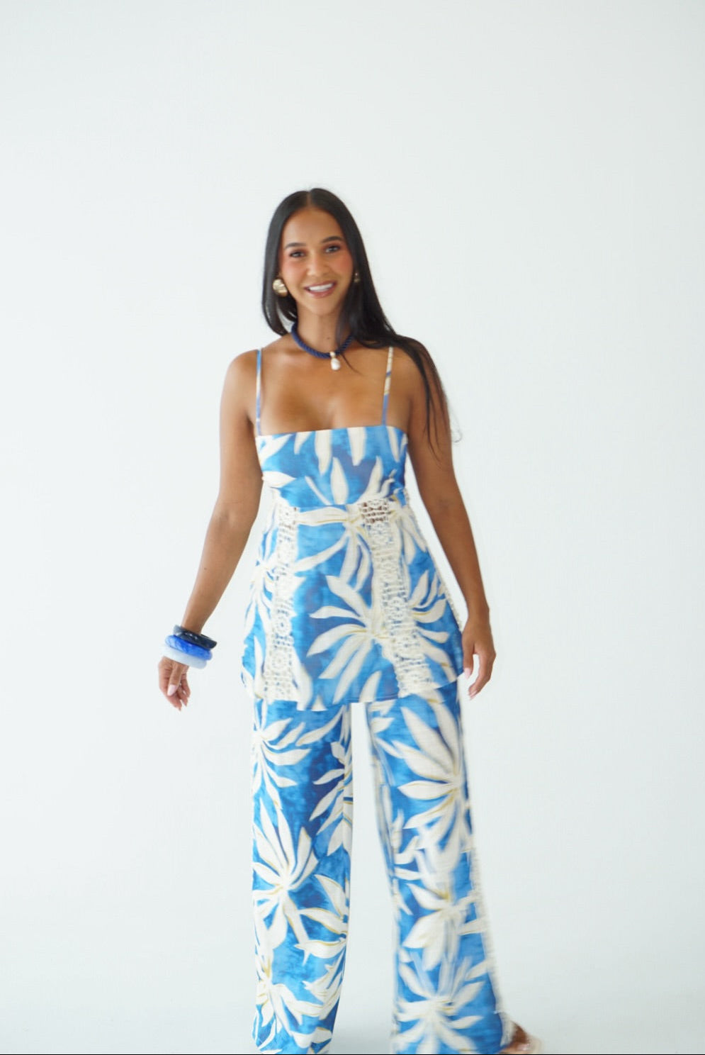 A woman modeling a blue and white tropical leaf print sleeveless jumpsuit with crochet detailing on the bodice, set against a plain white studio background.